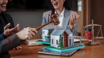 Real estate auction concept: gavel striking on model house atop laptop, with negotiators and scales of justice in background, symbolizing property law, sale, and contract agreement. © NanSan