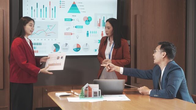 Business meeting presentation with three office professionals discussing charts on a large screen, laptop on table, modern conference room, teamwork and data analysis concept.