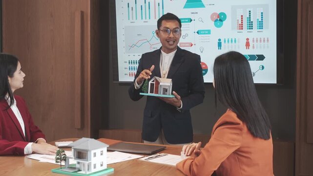 Business meeting with real estate agent presenting house model to clients in conference room, with charts on screen, modern office setting, teamwork and planning concept.