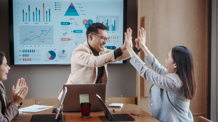 Naklejka premium Business team in modern meeting room celebrating with a high five during data presentation on large screen with charts and graphs, laptops on table, positive teamwork vibe.