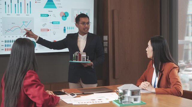 Business meeting presentation with man pointing at analytics screen while colleagues review model houses, discussing real estate investment strategy in modern office.