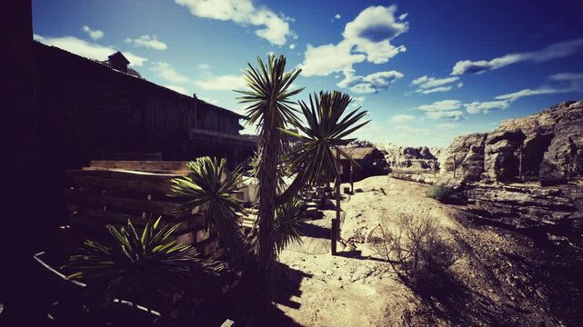 A stark desert landscape features tall, spiky plants next to wooden structures under a bright blue sky. Rocky formations rise in the background, showcasing the rugged beauty of the area.