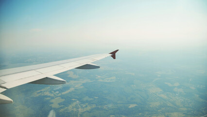 Fototapeta premium Airplane wing hovering over verdant countryside, revealing patchwork fields, rustic villages, and serpentine roads during sunny aerial journey