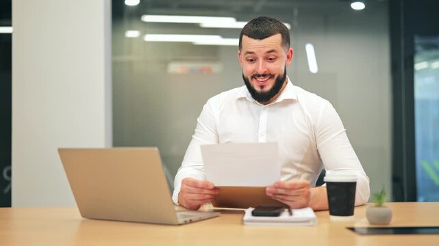 Excited bearded male executive in white shirt showing surprise and joy while reading documents. Businessman happy about unexpected positive results or profit in modern office workspace.