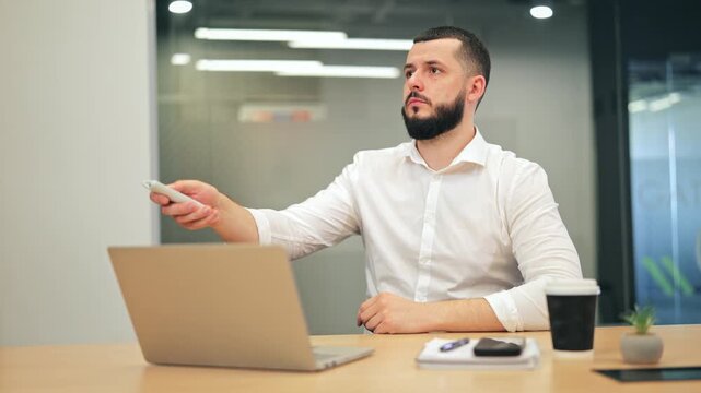 Bearded man in white shirt feeling cold at office desk, rubbing his arms and shoulders. Male executive using air conditioner remote control to increase heating in workspace.