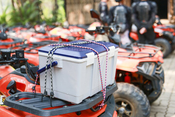 White cooler secured with strap on front rack of an orange all terrain vehicle stands ready for an exciting outdoor adventure alongside parked fleet of quads with riders © CoreRock