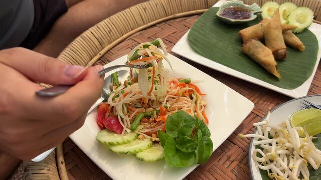 A person enjoys a delicious plate of authentic Thai Som Tum (Papaya Salad) garnished with fresh vegetables. Spring rolls and bean sprouts are also visible on a traditional woven tray.