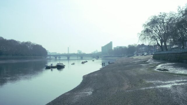 Cinematic aerial drone shot flying low over the River Thames, slowly approaching Putney Bridge in the distance. Calm, still water reflecting a soft hazy sky, creating a muted atmospheric tone. On the