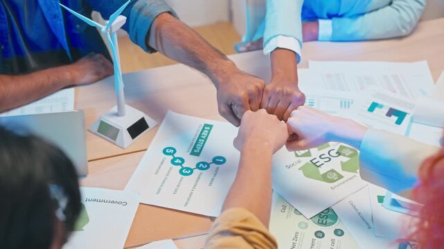 Multicultural colleagues bumping fists together over documents with ESG icons and a wind turbine model, celebrating successful sustainable business strategy.