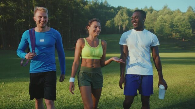 Laughing friends walk after exercising outside, Casual friends walk in sunlight with water bottles and towels