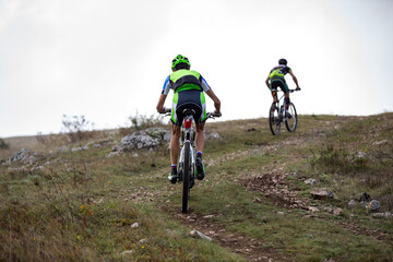 Fototapeta premium Two mountain bikers climbing grassy hill on trail under overcast sky