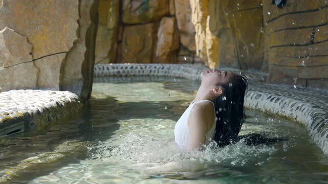 Beautiful young woman relaxing in a natural hot spring spa, swing long hair enjoys traditional onsen in open hot thermal swimming pool at sunrise for wellness travel and spa tourism industries