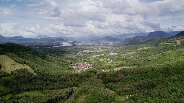 Slow hovering sideward motion flying view of mountainous tropical landscape, with the Mekong River in the distance, with sunny cloudy hazy environment in the background.