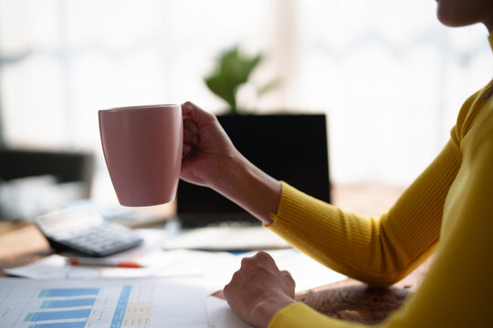 Woman holding coffee mug, taking a brief workday break at a desk scattered with financial reports, calculator and charts, pausing to relax and refocus amid paperwork
