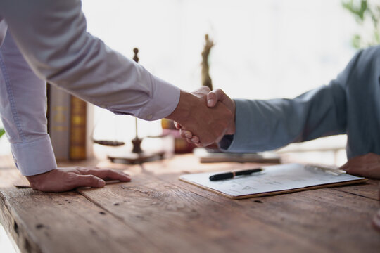 Two business professionals in formal attire shaking hands over a wooden desk with legal scales and a contract, symbolizing agreement, trust, and successful collaboration