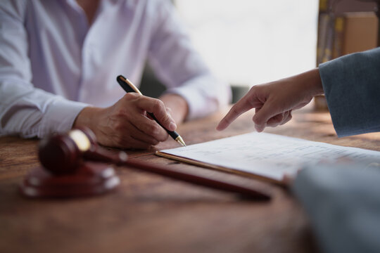 Client's hand signing a document with a pen during a legal consultation, while a lawyer points to a specific clause, with a gavel on the table symbolizing law and justice
