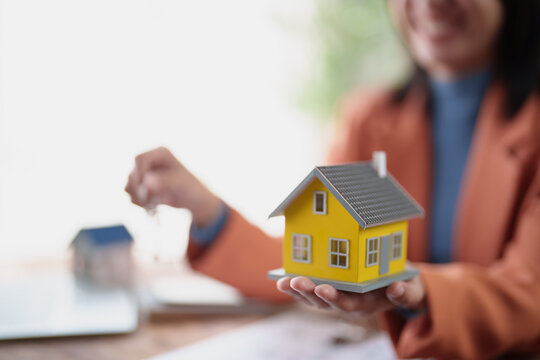 Real estate agent smiling while presenting a miniature house model and property keys, symbolizing new home ownership, investment, and successful property transactions