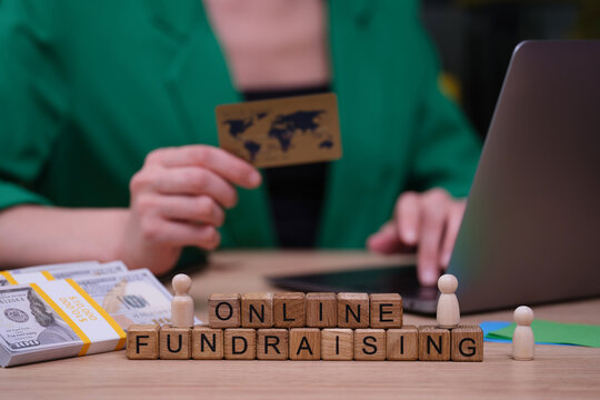 A person engages in online fundraising while holding a credit card, surrounded by stacks of cash and wooden blocks spelling out online fundraising.