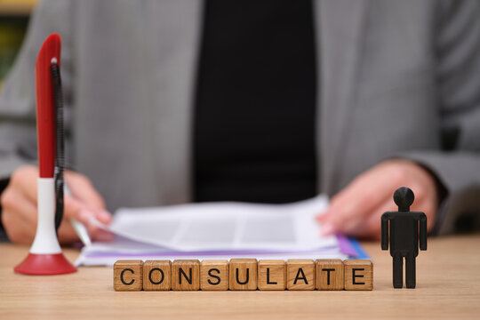 A person reviews important consulate documents at a desk, showcasing clear organizational tools. Wooden blocks spell out consulate beside a small figure, indicating an official process.
