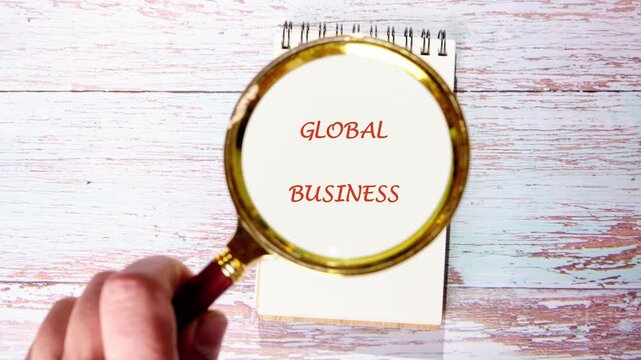 A hand focuses a golden magnifying glass on Global Business written on a cream notebook on a light wooden table