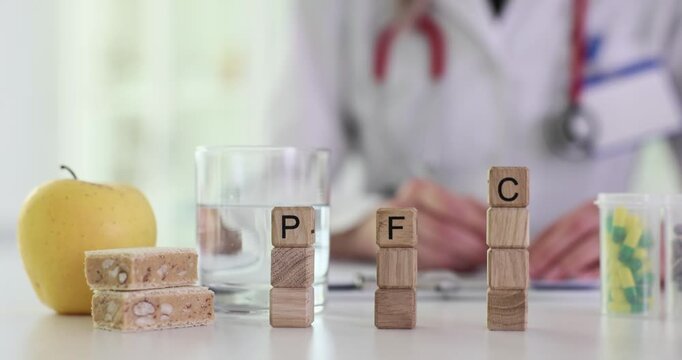 Wooden blocks show letters PFC near pills and apple on medical desk. Doctor writes notes behind blocks arranging items for nutrition plan for patient