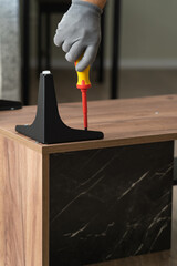 Hands assemble wooden nightstand using screwdriver. Installing metal furniture legs on coffee table. Process of assembling furniture