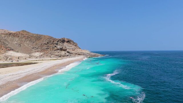 Aerial view of turquoise sea water meeting a white sand beach and rugged mountain cliffs along the coastline of Saudi Arabia.