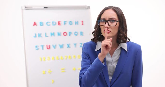 Teacher woman raises finger to lips demanding silence near alphabet board. Strict educator watches students enforcing classroom discipline slow motion