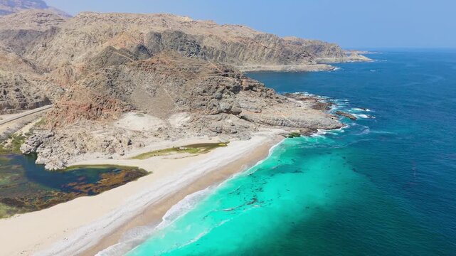 Aerial view of white sand beaches meeting turquoise waters alongside rugged mountain ranges and a coastal road in the Tabuk region, Saudi Arabia.