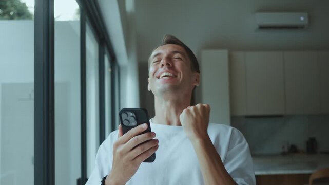 Man looks at smartphone and celebrates confirmation with confident yes gesture at home. This confirmation moment shows relief and success after approval payment or important message in daily life