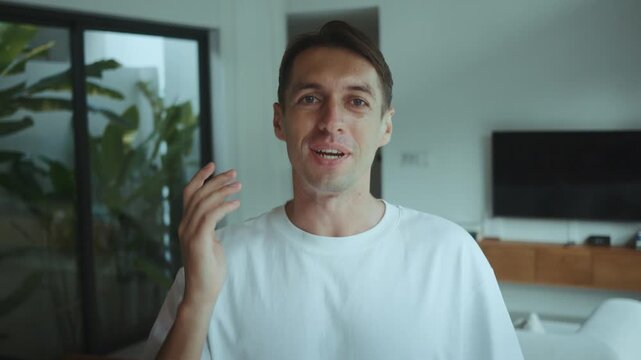 Smiling man looks at camera and greets audience with hand gesture while speaking emotionally This friendly vlogger talks directly to viewers creating personal connection online at home