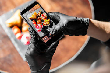 High-angle shot of a person wearing black nitrile gloves, holding a smartphone to take a photo of various gourmet appetizers and finger foods on a dark platter at a restaurant. © pavel siamionov