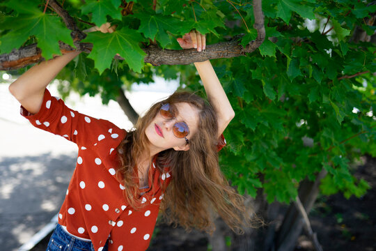 Woman with curly hair in red blouse and sunglasses posing under tree