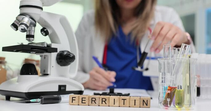 Wooden blocks spell word Ferritin near microscope and test tubes. Lab specialist woman pipettes blood sample writing note for iron evaluation