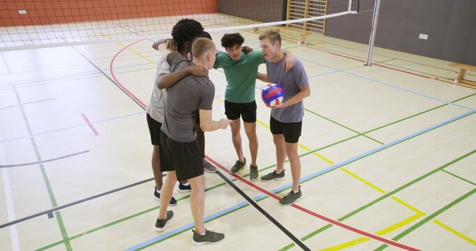 Diverse male teammates linking arms, stacking hands at center court with volleyball after point