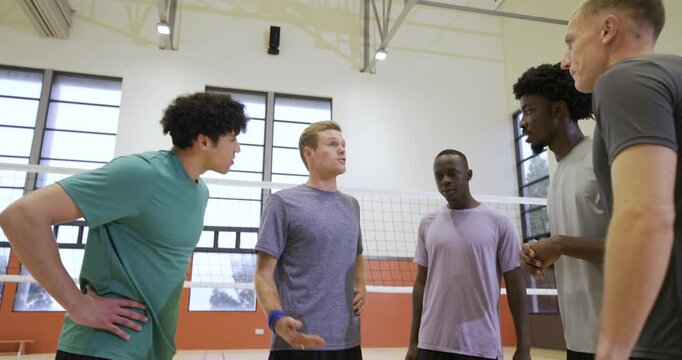 Five male teammates talking near volleyball net at gym, gray shirt initiating roles stacking hands