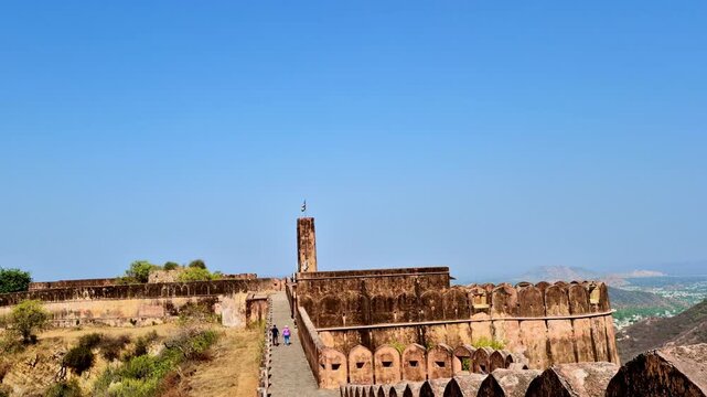 A bright daytime  showing the ramparts of Jaigarh Fort with stone walls, watchtower with Indian flag, and visitors walking along the fort pathway overlooking the Aravalli hills.