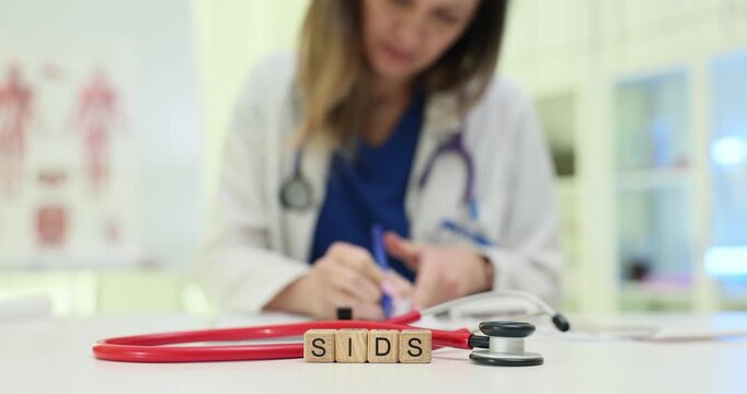 Wooden blocks spell abbreviation SIDS near stethoscope and clipboard. Doctor woman writes case notes checking infant risk factors during appointment