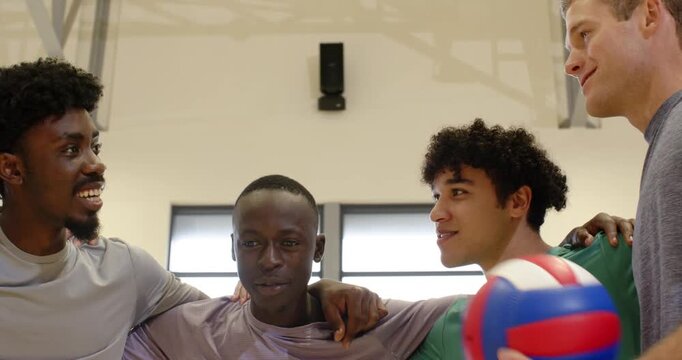 Diverse men huddling, linking arms, stacking hands, cheering before match at gym with volleyball