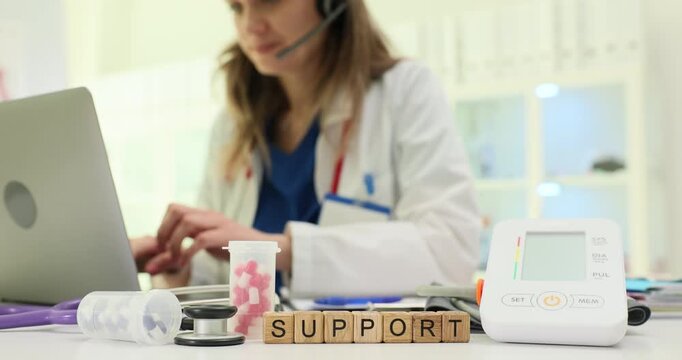 Wooden blocks spell word Support near laptop and medical monitor on desk. Doctor woman types case notes coordinating remote follow up for patient