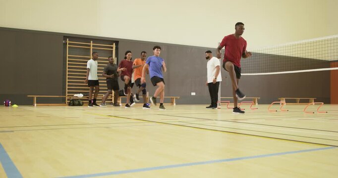 Diverse male team after coach starting timer doing high-knee drills over hurdles for agility in gym