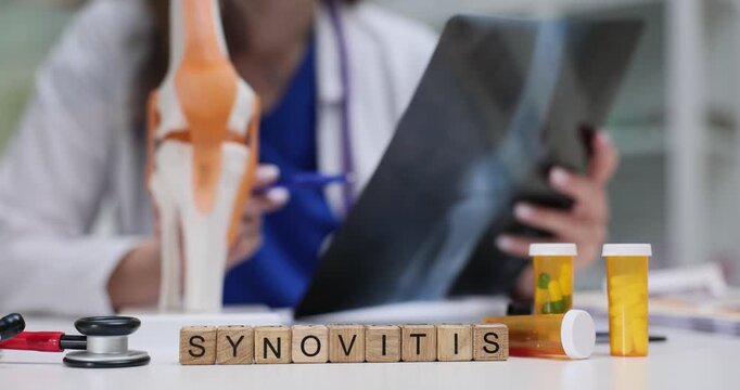 Woman doctor studies X-ray image of kneecap to evaluate bone alignment and joint structure. Wooden cubes neatly arranged to form word Synovitis