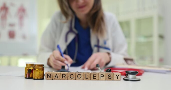 Woman doctor documents observations in patient record. Wooden cubes arranged on table to form term Narcolepsy placed near tablets and stethoscope
