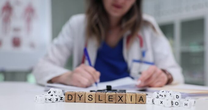 Miniature wooden cubes carefully arranged on table form word Dyslexia. Woman doctor writes detailed recommendations on sheet of paper for patient