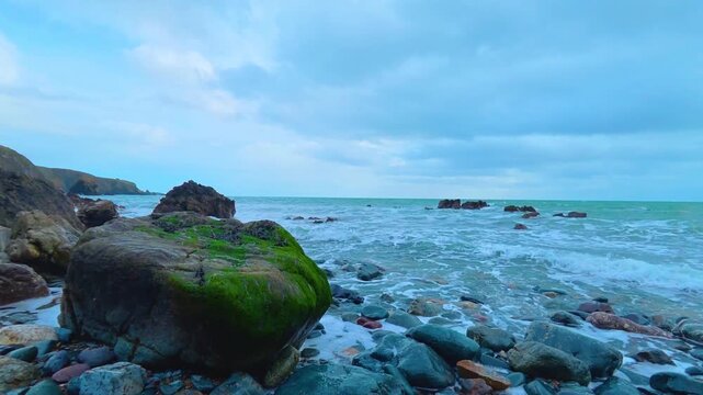 Timelapse Copper Coast Waterford Ireland large green lichen covered bolder multi coloured small rocks gentle lapping waves and slowly moving clouds on a spring morning