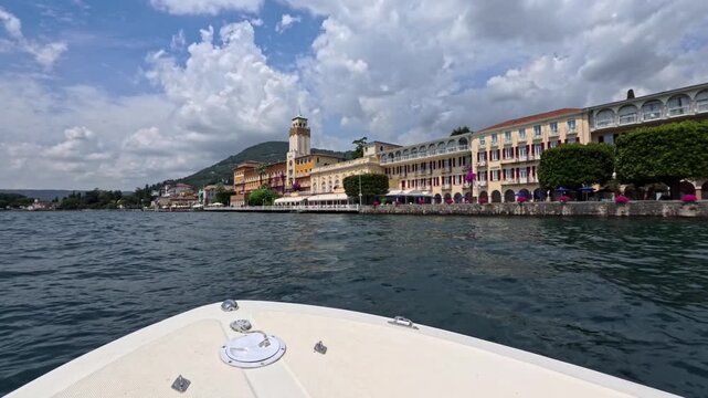 Boat view of Gardone Riviera waterfront on Lake Garda, motorboat glides past pastel hotels and arcades with flowered promenade under towering summer clouds in Italy