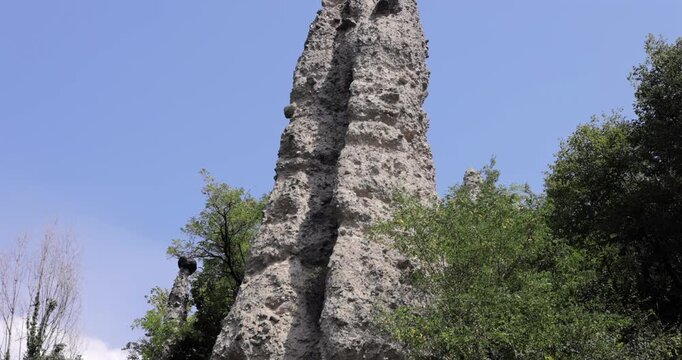 Close up earth pyramid hoodoo (Piramidi di Zone) at Lake Iseo Lombardy Italy, camera tilts from rough clay base up to a dark boulder capstone framed by summer trees and blue sky