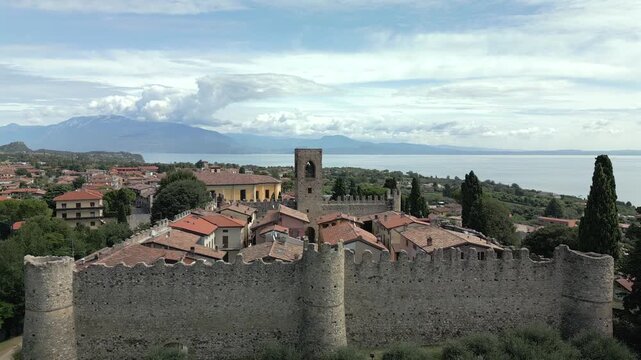 Drone boom down with tilt up over Castello di Moniga del Garda, stone battlements frame red roofs as Lake Garda and hazy mountains fill the bright sky above calm water