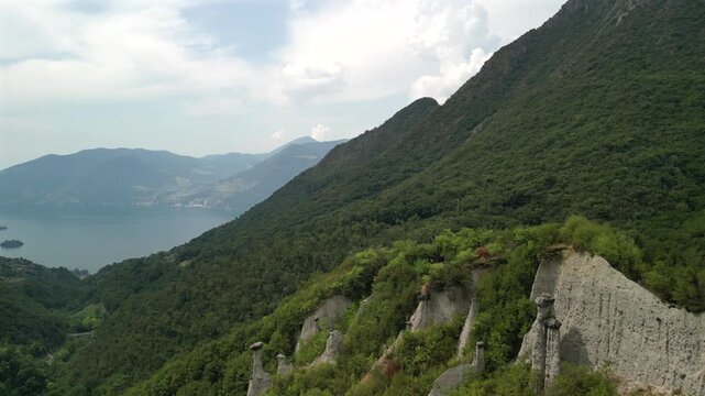 Drone flight over Zone earth pyramids (Piramidi di Zone) near Lake Iseo in Lombardy Italy, smooth forward glide reveals hoodoo pillars and green slopes with the blue lake and mountains fading in haze