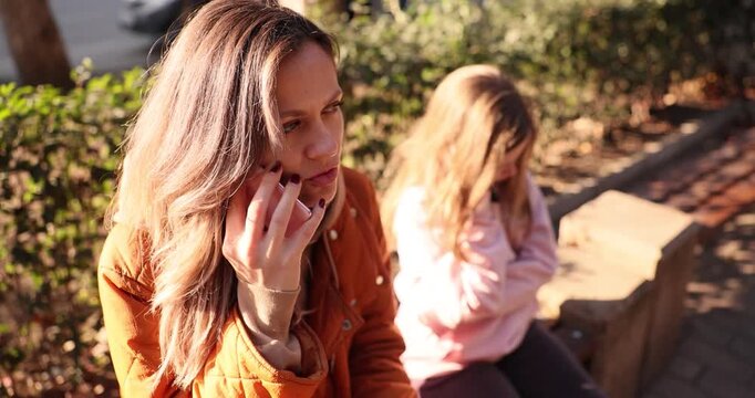 Worried mother talks on phone in sunny park at warm autumn light. Sad daughter sits near mother on bench looking down showing stressful family moment